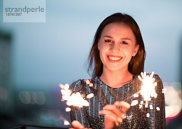 Entzückte junge Dame im Abendkleid  die lächelt und mit brennenden Wunderkerzen während einer nächtlichen Weihnachtsfeier winkt