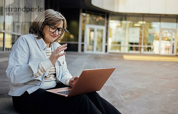 Glückliche Frau mittleren Alters  die einen Videoanruf auf dem Laptop tätigt  auf dem Bildschirm winkt und online mit Menschen aus der Ferne plaudert. Lächelnde grauhaarige ältere Frau winkt hallo  während auf einem Videoanruf auf der Treppe im Freien sitzen