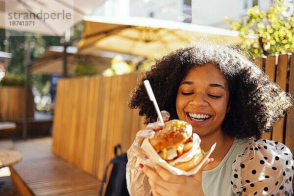 Junges afrikanisches amerikanisches Mädchen ißt klassischen Burger und trinkt Limonade in der Stadt. Outdoor Frau trägt Sommerbluse mit Tupfen eatting leckeren Burger beim Sitzen auf der Terrasse. Frau in blau Verkostung Sandwich mit freudigen Blick bewundern sonnigen w