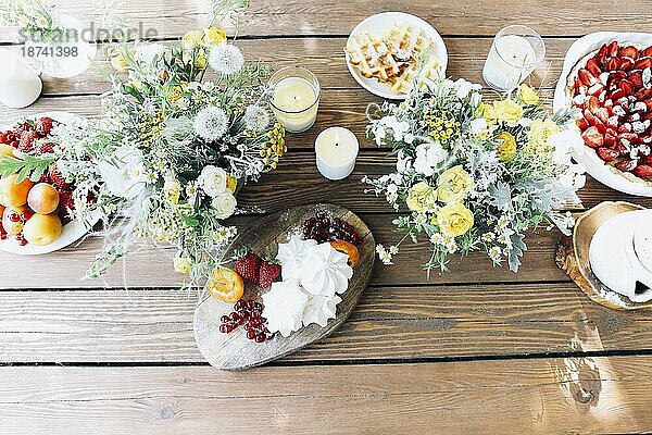 Draufsicht auf leckeren Erdbeerkuchen und hausgemachte Waffeln auf einem Holztisch mit Blumensträußen auf dem Lande an einem sonnigen Tag