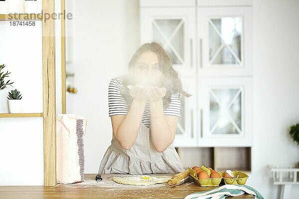 Junge Frau Hausfrau in Schürze bläst Mehl während der Herstellung von Teig und Kochen in hellen Küche zu Hause  spielerisch weiblichen Spaß haben  während Backen Kuchen für Familie am Wochenende