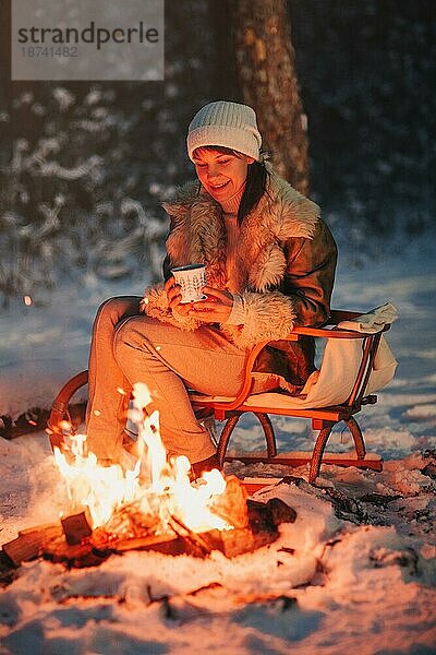 Glückliche junge Frau voller Ruhe und Gelassenheit in warmer Strickmütze und Jacke sitzt auf einem Holzschlitten am Lagerfeuer mit einer Tasse heißem Tee in der Hand  umgeben von einem schneebedeckten Winterwald