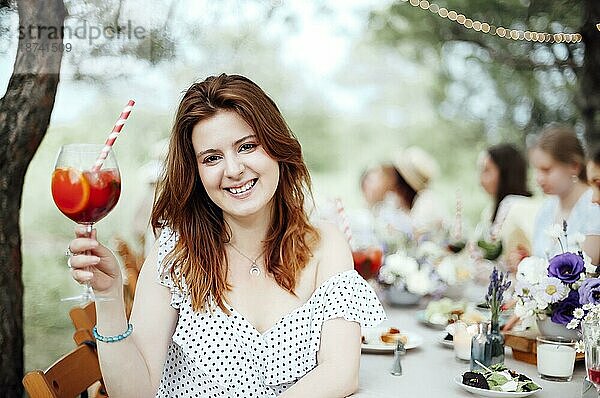 Glückliche junge Frau mit Alkoholcocktail  die lächelt und einen Toast ausspricht  während sie bei einem Bankett im Garten am Tisch sitzt