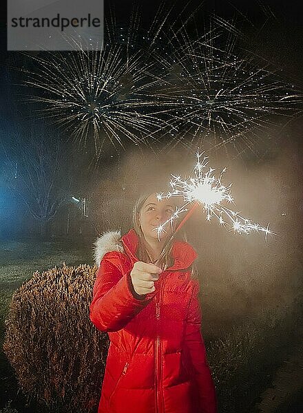 Glückliches Teenagermädchen das eine Wunderkerze in der Hand hält  während es im Freien in der Dämmerung während der Winterferien Neujahr feiert  Feuerwerk am Himmel auf dem Hintergrund