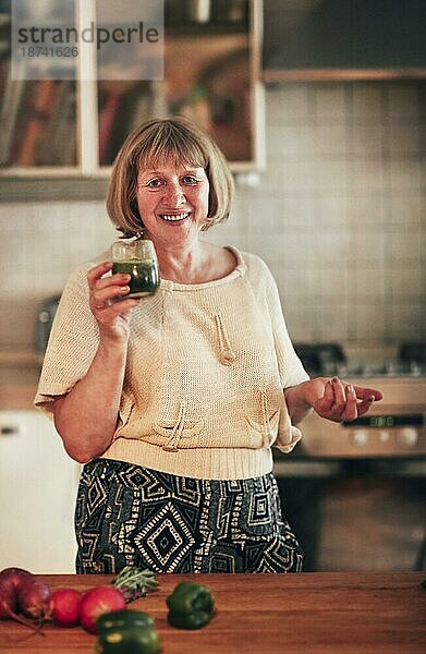 Glückliche Frau mittleren Alters mit frischem Gemüsesmoothie  die lächelt und in die Kamera schaut  während sie in der Küche zu Hause steht