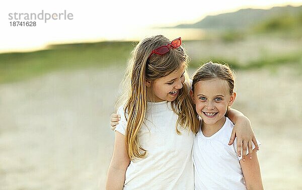 Fröhliche kleine Schwestern  die sich glücklich umarmen  während sie gemeinsam am sandigen Meeresufer bei Sonnenuntergang stehen