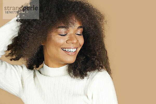 Porträt einer wunderschönen  fröhlichen  afrikanisch amerikanischen jungen Frau mit lockigem Haar  die lächelnd vor einem beigen Studiohintergrund steht  eine positive schwarze Frau mit gesunder Haut und strahlendem Lächeln