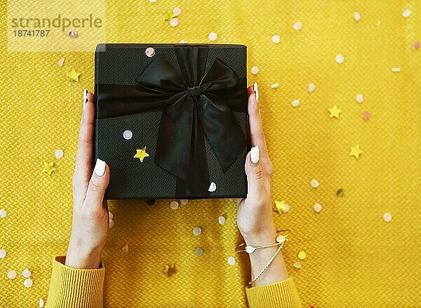 Neujahr oder Weihnachten flach legen. Draufsicht Xmas Feiertag Feier dekorative schwarze Geschenkbox mit schwarzer Schleife  hell funkelt auf dem gelben Hintergrund mit Kopie Raum. Grußkarte Ihr Text Design