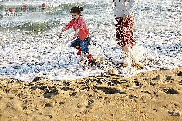 Junge liebende Mutter mit lächelnder Tochter  die ihr am sonnigen Strand entgegenläuft  glückliche Mutter umarmt kleines Mädchen und genießt die gemeinsame Zeit am Sommerstrand. Rückansicht