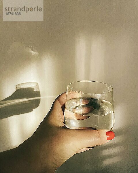 Morgendliche Flüssigkeitszufuhr. Weibliche Hand hält Glas reines sauberes Mineralwasser gegen beige Wand mit Schatten  beschnitten Schuss der Frau verhindert Organismus vor Austrocknung. Gesundheitspflege und Entgiftung Konzept