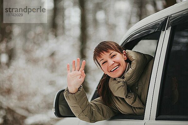 Fröhliche Frau lächelt und schaut in die Kamera  während sie ein modernes Fahrzeug auf einem unscharfen Hintergrund einer verschneiten Landschaft fährt