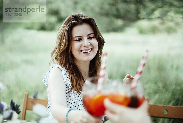 Glückliche junge Frau mit Alkoholcocktail  die lächelt und einen Toast ausspricht  während sie bei einem Bankett im Garten am Tisch sitzt