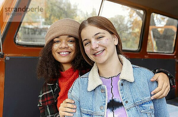 Picknick im Herbst Natur. Zwei glückliche multirassische Freundinnen sitzen auf dem Kofferraum eines Autos im Wald  heben die Arme und lächeln in die Kamera während eines abenteuerlichen Roadtrips in der Herbstsaison  lächelnde Freundinnen im Minivan