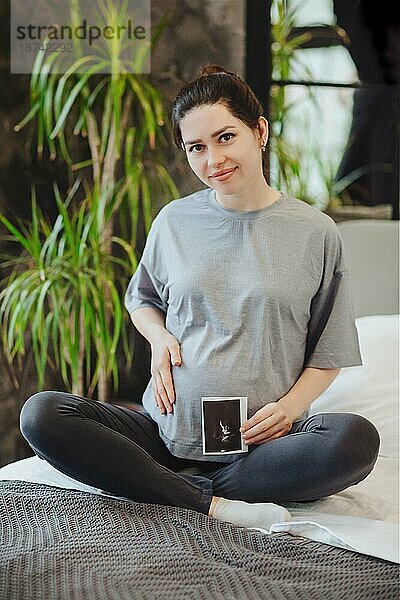 Schwangere Frau hält Schwangerschaft Ultraschall beim Sitzen in Lotuspose auf dem Bett zu Hause  werdende Mutter mit Baby Sonogramm. Schwangerschaft  Mutterschaft und vorgeburtliche Betreuung