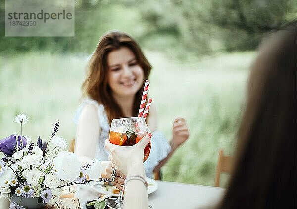 Glückliche junge Frau mit Alkoholcocktail  die lächelt und einen Toast ausspricht  während sie bei einem Bankett im Garten am Tisch sitzt