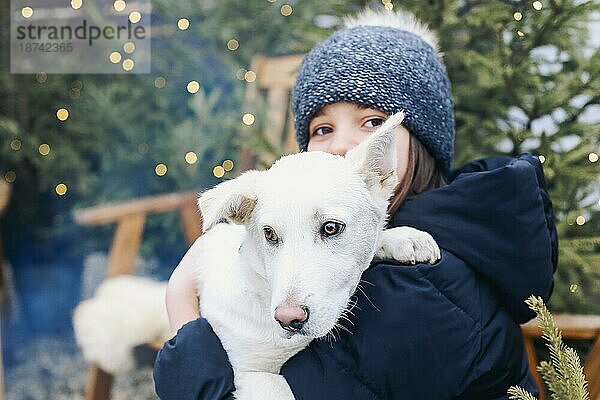 Lächelndes Teenagermädchen in warmer Kleidung  das ein süßes weißes Hündchen in den Armen hält  während es in einem Hinterhof mit Nadelbäumen sitzt  den Hund umarmt und das Winterwetter in Gesellschaft seines besten Freundes genießt