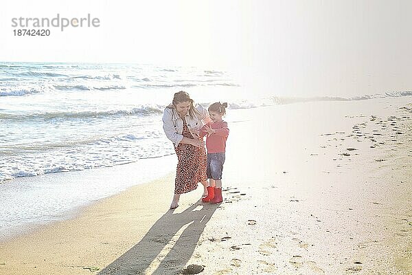 Glückliche Familie von zwei  lächelnde Mutter und niedliche kleine Tochter  zu Fuß entlang sonnigen Sandstrand  verbringen Zeit am Meer zusammen. Atmosphärisches Lifestylefoto von aktiven Eltern  die mit Kindern unterwegs sind