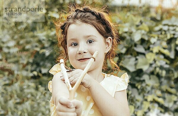 Nettes freches rothaariges kleines Mädchen mit geflochtenem Haar  das mit einer Steinschleuder auf die Kamera zielt  während es einen Sommertag im Garten verbringt