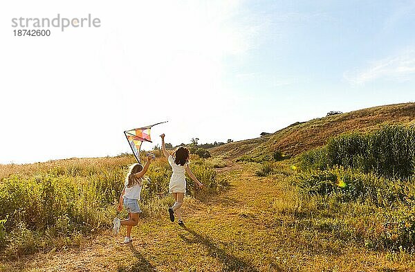 Unbeschwerte Frau und Teenagermädchen die an einem Sommerwochenende auf einem Feld entlanglaufen und mit einem Drachen spielen