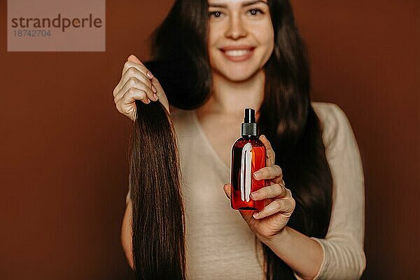 Studioporträt einer glücklichen  zufriedenen Frau  die ihr langes  gesundes  braunes Haar hält und in die Kamera zeigt  daß sie Shampoo oder Spülung benutzt  eine Frau  die natürliche  parabenfreie Haarpflegeprodukte verwendet  selektiver Fokus