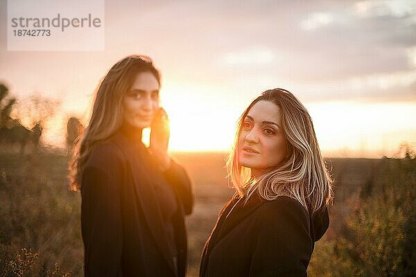 Zwei glückliche Frauen  die sich an den Händen halten  spazieren durch ein herbstliches Feld bei Sonnenuntergang und lächeln  sorglose beste Freundinnen  die den Abendspaziergang auf einer Wiese auf dem Lande genießen