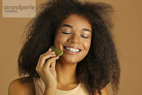 Gorgeous dunkelhäutigen Modell weint sanft Gua sha Stein bis Wangenknochen massiert ihr Gesicht zu helfen  Flüssigkeiten abfließen und fördert die Durchblutung  lächelt mit geschlossenen Augen isoliert über beige Hintergrund