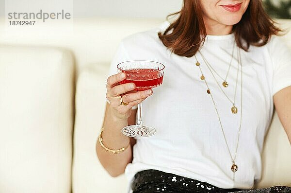 Abgeschnittene Aufnahme einer jungen lächelnden Frau  die ein Cocktailglas mit rotem Alkohol in der Hand hält und zu Hause auf dem Sofa sitzt. Sie trägt modische Kleidung und Accessoires und genießt eine Party mit Freunden im Haus