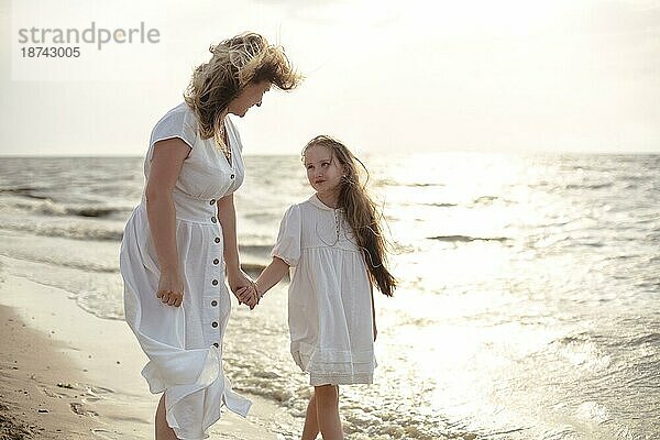 Ganzkörper fröhliche Frau in weißem Kleid umarmt Mädchen  während stehend am Strand in der Nähe winken Meer am Morgen