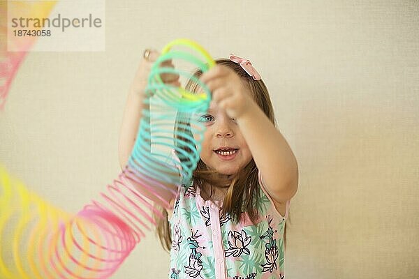 Cute adorable kleines Mädchen spielt mit Kunststoff Regenbogen magische Feder  hält bouncy dehnbar Spielzeug und lächelt in die Kamera. Glückliches Kind vorbeige Hintergrund genießen Freizeit zu Hause