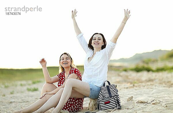 Seitenansicht von begeisterten jungen Freundinnen  die im Sand sitzen und den Sommer genießen  während sie zusammen am Meer Urlaub machen