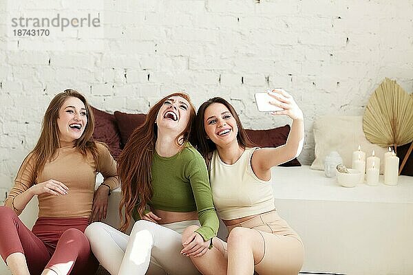 Optimistische junge Frauen in legerer Kleidung lächelnd und nehmen selfie beim Sitzen gegen weiße Backsteinwand zu Hause