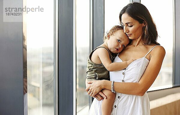 Mutter sein. Junge schöne Frau Mutter hält niedlichen kleinen Jungen  während durch Fenster zu Hause stehen. Frau in weißem Kleid kuscheln ihren kleinen Sohn Säugling und zum Ausdruck bringen Liebe und Sorgfalt