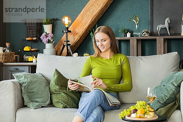 Junge blonde Frau  die eine interessante Zeitschrift liest  während sie auf einer bequemen Couch in der Nähe eines Tisches mit Obst und Wein an einem Wochenendtag zu Hause sitzt