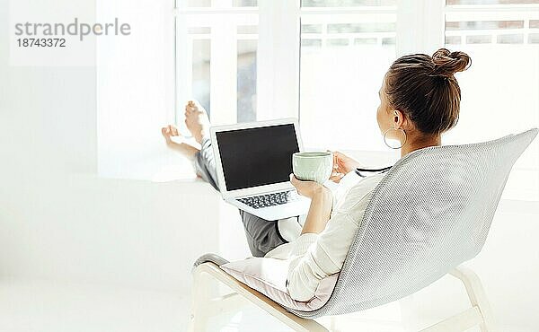 Barfüßige junge Frau mit Händen hinter dem Kopf  die auf einem Stuhl sitzt und sich mit geschlossenen Augen entspannt  nachdem sie einen Laptop in einem hellen Raum zu Hause benutzt hat