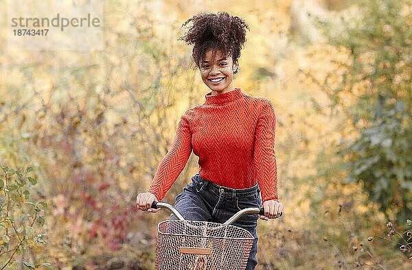 Junge glückliche lächelnde afroamerikanische Frau beim Fahrradfahren im Herbstwald  aktive positive gemischtrassige Frau in Freizeitkleidung  die in die Kamera lächelt  während sie mit dem Stadtrad in der herbstlichen Natur steht