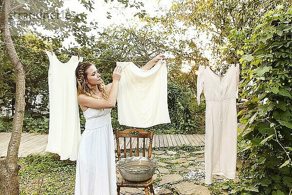 Junge Frau hängt Wäsche im Freien auf. Nettes Mädchen im Kleid wäscht weiße Wäsche in einem Metallbecken im Hinterhof  hängt Wäsche auf Wäscheleine und lässt sie im Garten trocknen  trocknet Kleidung im Freien im Wind