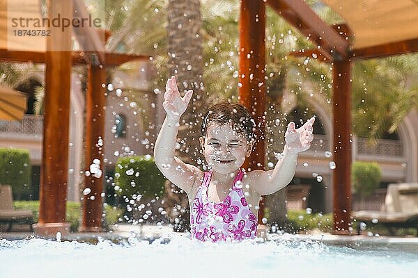 Adorable Mädchen im Badeanzug spritzt Wasser und haben Spaß im Schwimmbad im Sommer beim Blick in die Kamera