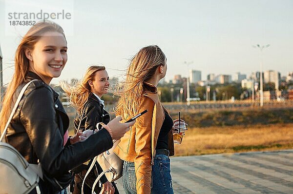 Glückliche junge Frauen mit Getränken zum Mitnehmen  die auf dem Bürgersteig spazieren gehen und an einem sonnigen Wochenendtag auf einer Straße in der Stadt lächeln