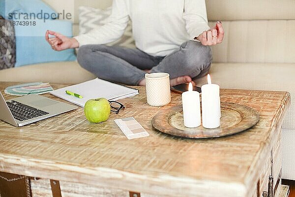 Crop junge Frau in Freizeitkleidung meditieren in Lotuspose beim Sitzen auf dem Sofa vor dem Tisch mit Laptop und brennenden Kerzen in gemütlichen Wohnzimmer zu Hause