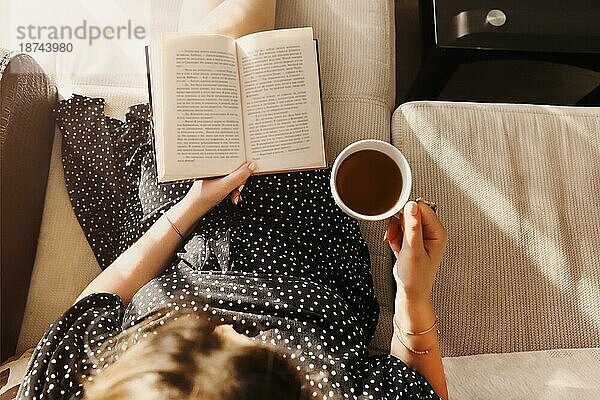 Foto von oben von junger Frau in legerem Kleid sitzt mit Buch und Tasse Tee auf gemütliche Couch zu Hause  entspannt glückliche Dame verbringen Freizeit in der Stille genießen Lesen und ihr Lieblingsgetränk