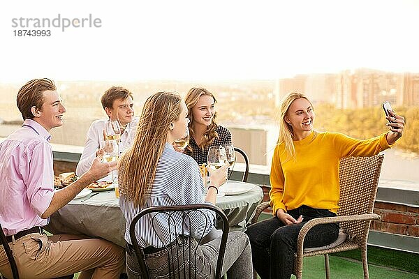 Gruppe von fünf fröhlichen jungen Menschen beim Abendessen im Restaurant  sitzen auf der Terrasse am Tisch und halten Gläser Wein in den Händen posieren für selfie auf Smartphone. Freundschaft und Versammlung Konzept