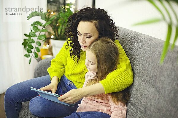 Mutter und kleines Mädchen Tochter beobachten Cartoons auf digitalen Tablet  während auf dem Sofa zu Hause sitzen  selektiven Fokus. Junge moderne Mutter und kleines Kind mit Gerät bei der Freizeitgestaltung zusammen