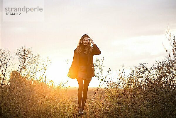 Junge glückliche kaukasische brünette Frau  die bei Sonnenuntergang über eine Wiese läuft und lächelt  verträumte schöne Frau im Abendsonnenlicht  die auf einem herbstlichen Feld auf dem Lande steht