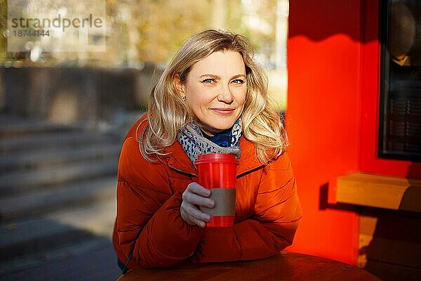 Erwachsene blonde Frau in roter Oberbekleidung  die lächelt und in die Kamera schaut  während sie an einem Tisch sitzt und ein Getränk zum Mitnehmen in einer Straßencafeteria genießt