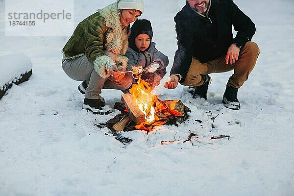 Fröhliche Eltern und Sohn genießen ein heißes Getränk  während sie sich abends am Lagerfeuer im Winterwald ausruhen