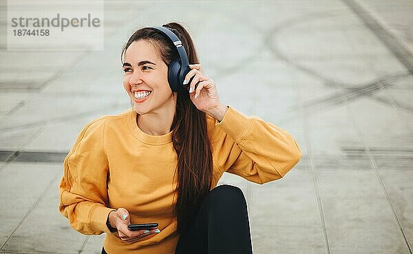 Außenaufnahme einer glücklichen jungen Frau mit Kopfhörern  die Musik auf ihrem Smartphone hört und lächelt  während sie auf der Straße in einem leeren Stadion sitzt  gekleidet in Freizeitkleidung