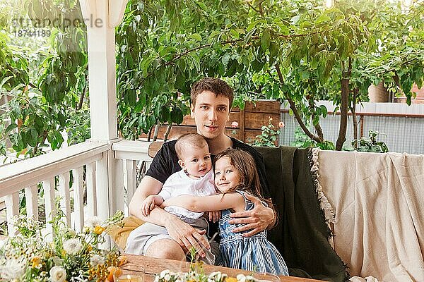 Ruhiger Mann  der auf einer Terrasse sitzt und einen kleinen Jungen und ein Mädchen umarmt  während er in die Kamera schaut und sich auf dem Land im Sommer entspannt