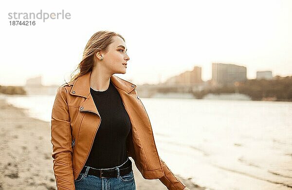 Glückliche junge Frau in modischer Lederjacke lächelt und schaut in die Kamera  während sie am Ufer eines Sees in einem windigen Abend steht