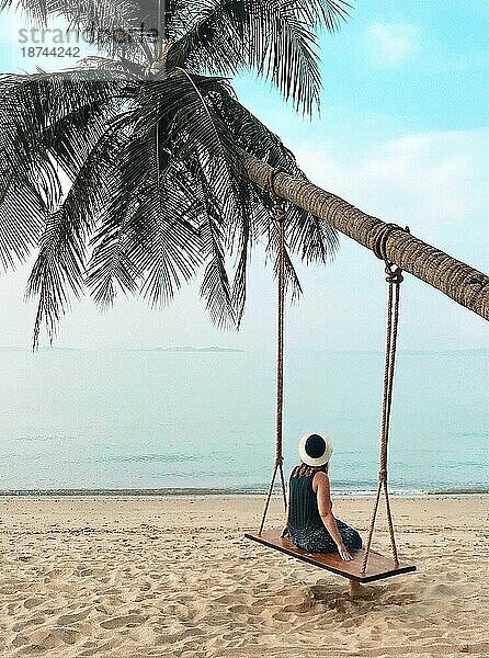 Rückansicht der Frau sitzt auf Schaukel hängen auf Palme und Blick auf ruhigen blaün Ozean  vertikale Schuss weibliche Entspannung auf Paradies Sandstrand und genießen Urlaub in tropischen Land