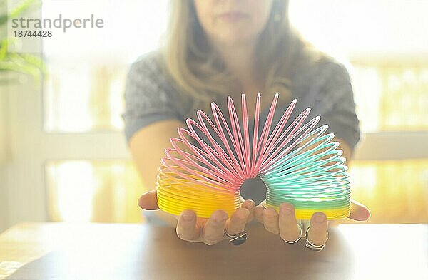 Ausgeschnittene Aufnahme einer Frau  die mit einer magischen Regenbogenfeder aus Plastik spielt  um Stress abzubauen. Eine Frau mittleren Alters hält ein elastisches  dehnbares Spielzeug in der Hand  während sie am Tisch in einem sonnigen Zimmer zu Hause sitzt. Selektiver Fokus